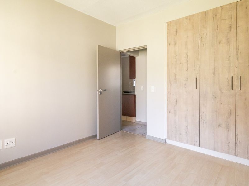 A clean, empty bedroom featuring light-toned laminate flooring and modern built-in wooden cupboards, with a view into the adjacent kitchen area.