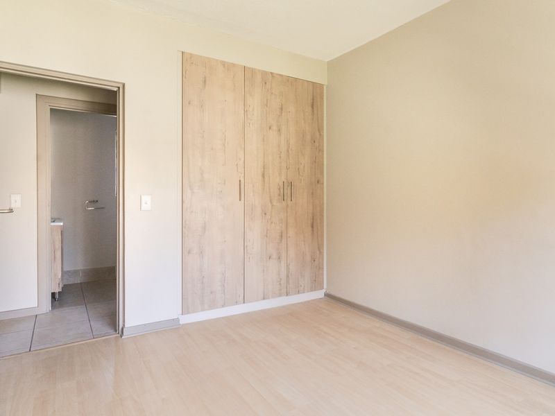 A bright, empty bedroom featuring modern light wood-look built-in cupboards, laminate flooring, and direct access to an en-suite bathroom.