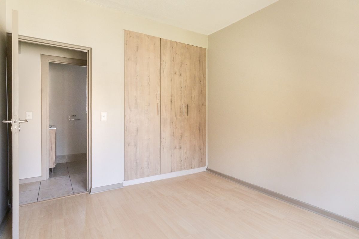 A bright, empty bedroom featuring modern light wood-look built-in cupboards, laminate flooring, and direct access to an en-suite bathroom.
