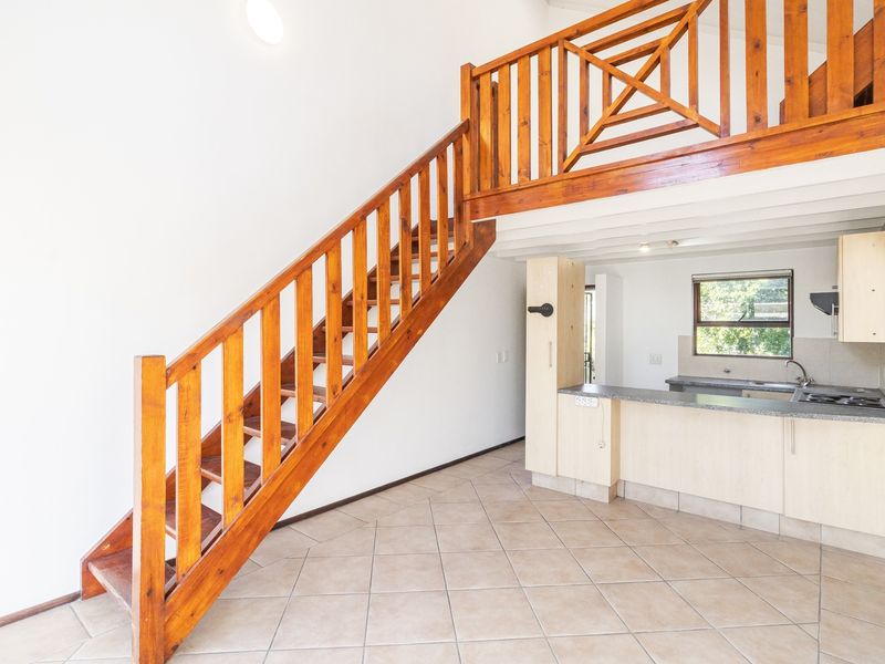 Open-plan living area featuring a wooden staircase leading to a mezzanine level, adjacent to a functional kitchen with built-in cabinetry and tiled flooring.