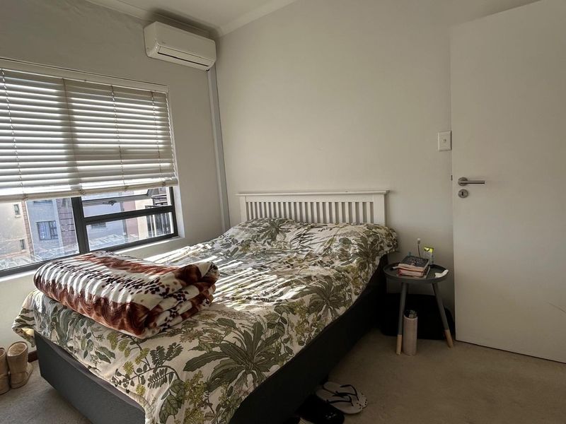 A functional bedroom featuring a wall-mounted split-system air conditioner, carpeted flooring, and a window fitted with venetian blinds.