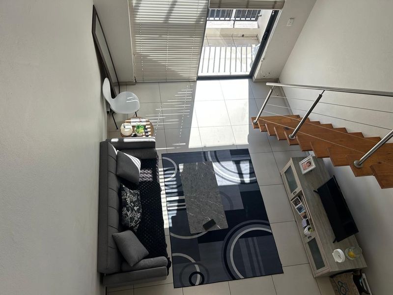A bright, open-plan living area featuring tiled floors, a wooden staircase with a stainless steel balustrade, and direct access to a balcony.