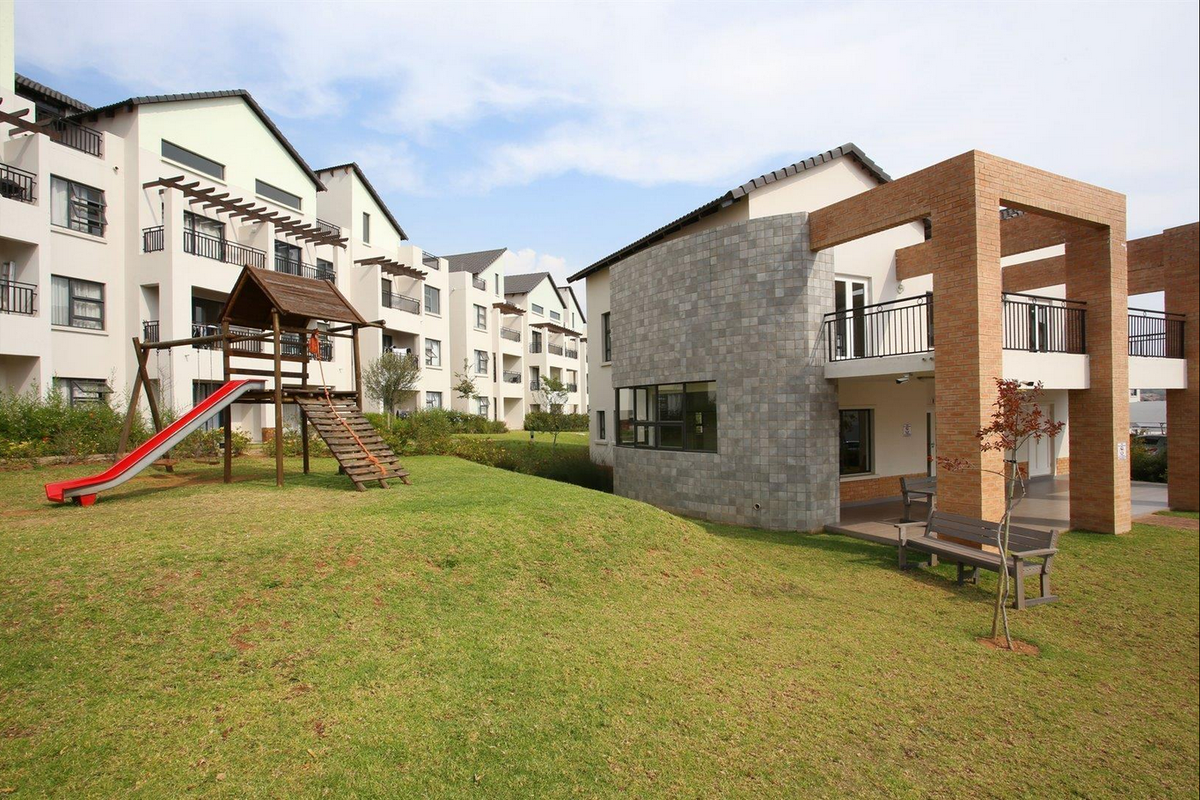 A well-maintained communal garden area featuring a lawn, playground equipment, and modern apartment building facades with brick and stone architectural details.