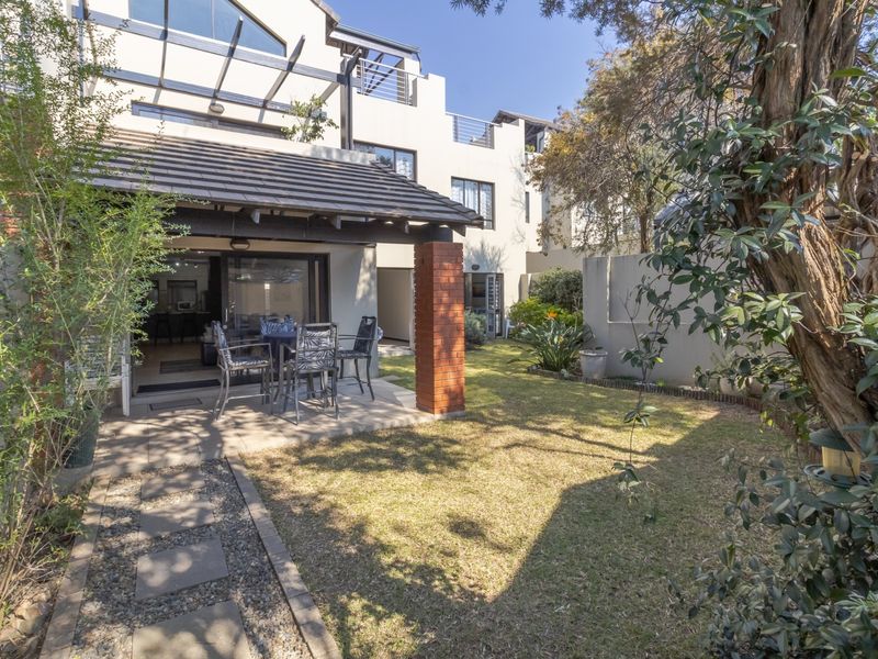 A private rear garden featuring a covered patio area with brick support pillars, paved pathways, and direct access to the apartment interior.