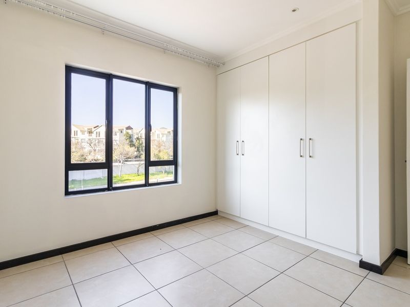 A bright, empty bedroom featuring neutral tiled flooring, built-in cupboards, and large aluminium-framed windows providing natural light.