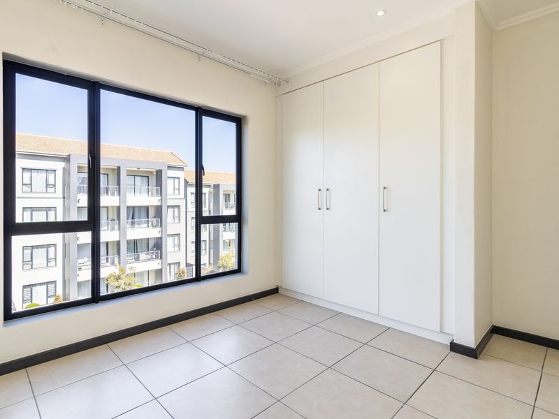A bright, empty bedroom featuring neutral tiled flooring, built-in cupboards, and large windows offering natural light and an urban view.