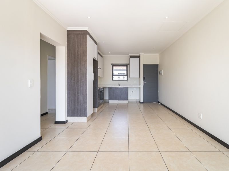 An open-plan living and kitchen area featuring neutral tiled flooring, modern built-in cabinetry, and integrated kitchen appliances in a clean, vacant space.