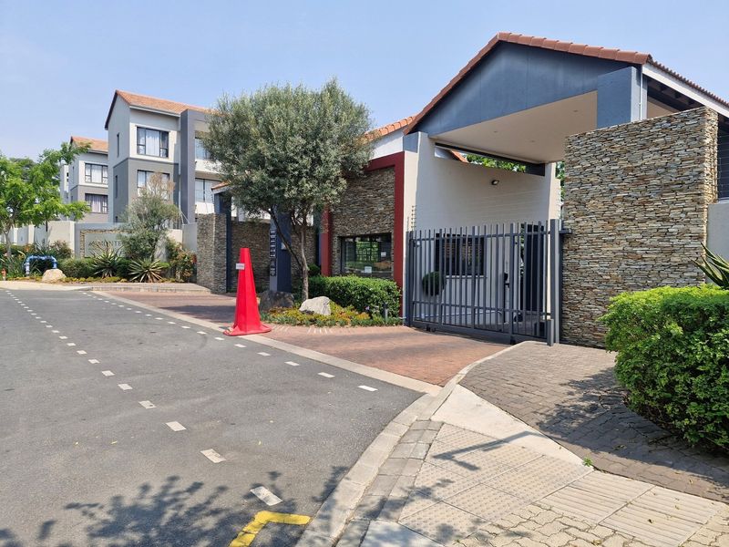 The entrance to the apartment complex features a secure gated access point with stone cladding, a guard house, and paved driveway.