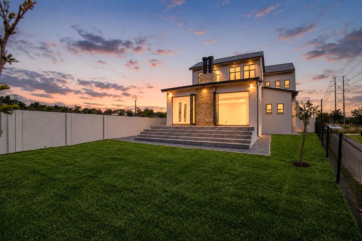 Rear view of a modern two-story house at dusk, featuring a stone-clad chimney, bi-fold doors opening to a patio, and a well-maintained lawn.