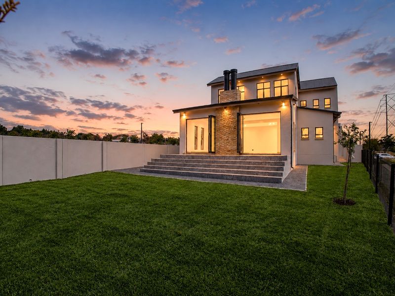 Rear view of a modern two-story house at dusk, featuring a stone-clad chimney, bi-fold doors opening to a patio, and a well-maintained lawn.