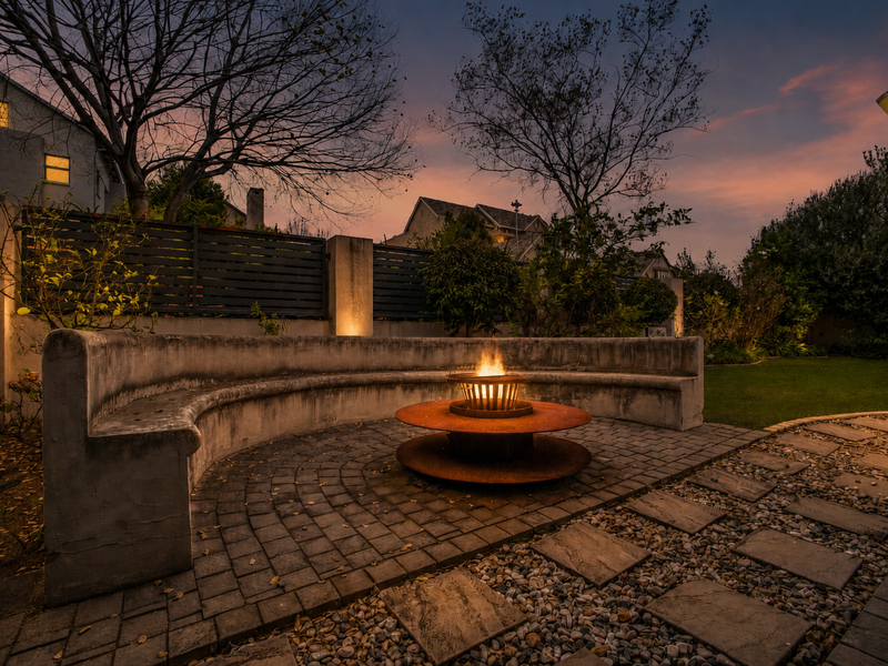 A beautifully landscaped rear garden featuring a paved patio area with a built-in concrete bench and a central fire pit, perfect for outdoor entertaining.