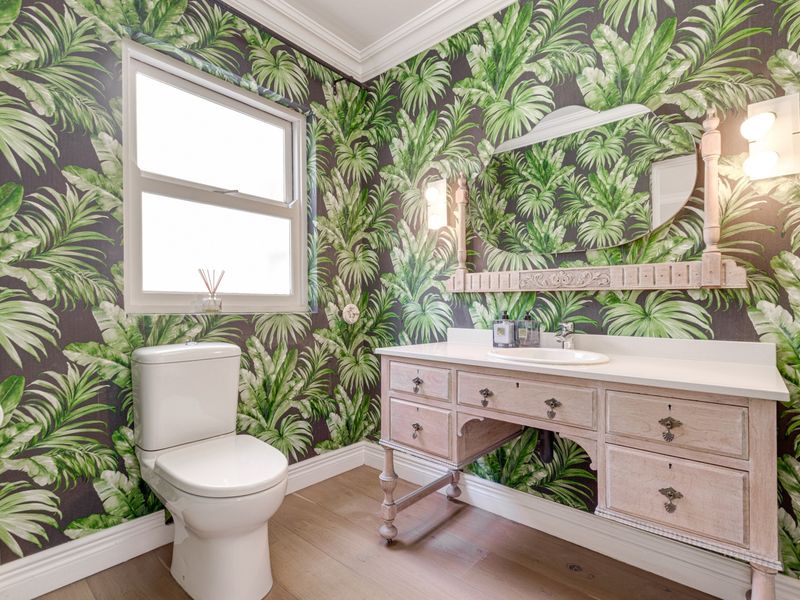 A well-maintained bathroom featuring bold tropical-patterned wallpaper, a classic vanity with integrated sink, and elegant wall sconces.
