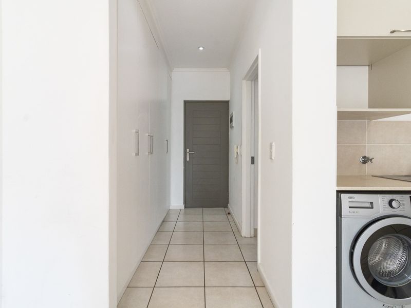 A clean, neutral hallway featuring tiled flooring, ample built-in cupboard storage, and a direct view into an adjacent kitchen area with integrated laundry plumbing.