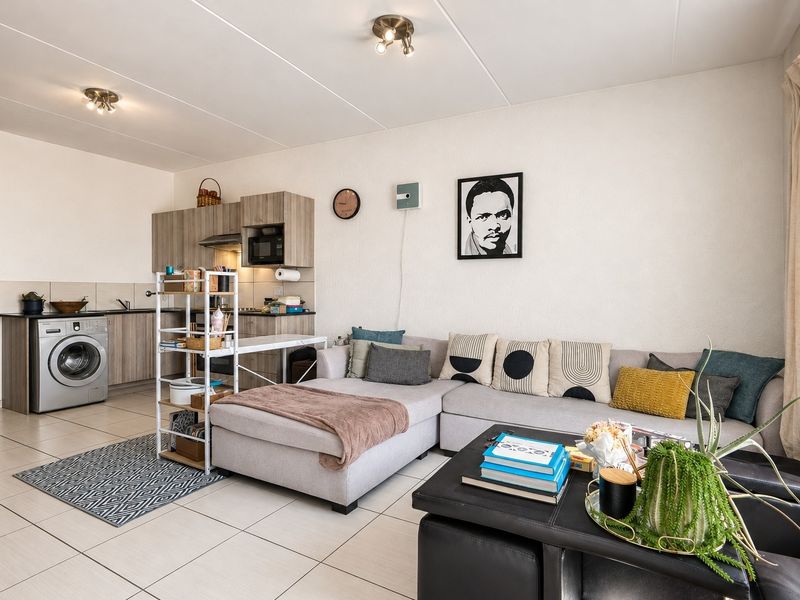 Open-plan living area featuring a compact kitchen with built-in cabinetry, integrated laundry appliances, and neutral tiled flooring throughout.