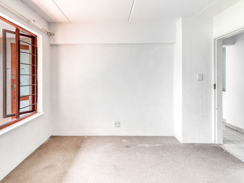 An empty bedroom featuring carpeted flooring, a large casement window with garden views, and direct access to an adjacent tiled hallway.