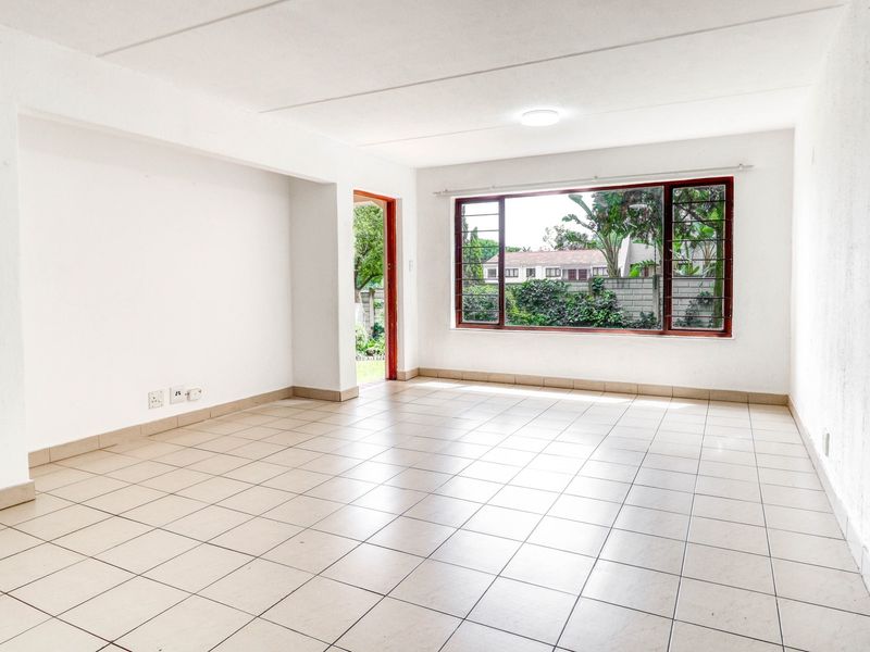 A bright, empty living room featuring neutral tiled flooring, large windows with security bars, and direct access to an outdoor area.