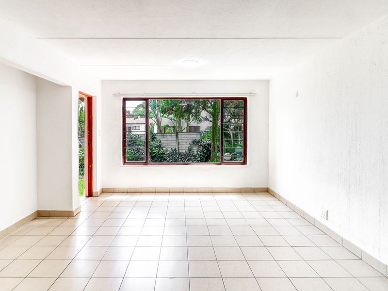 A bright, empty living room featuring neutral tiled flooring, white walls, and a large window providing natural light and garden views.