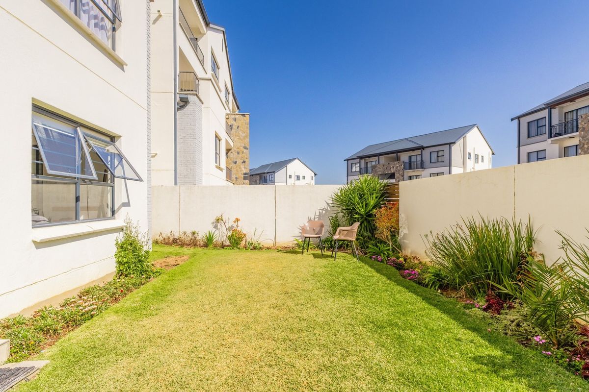 A private rear garden area featuring a well-maintained lawn, landscaped garden beds, and a secure boundary wall, adjacent to the apartment building.