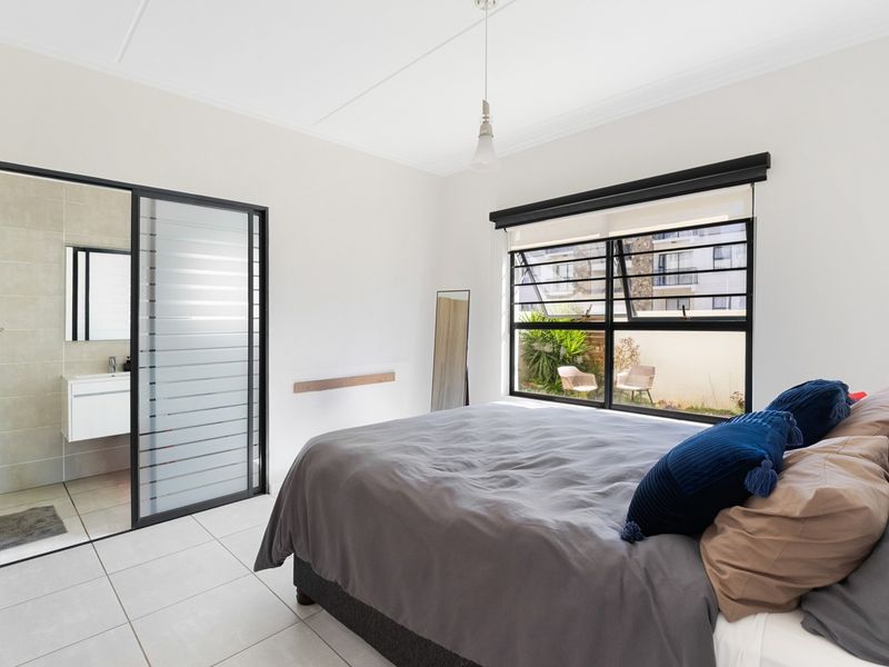 A bright bedroom featuring tiled flooring, a large window with black frames, and a modern en-suite bathroom accessed via a sliding glass door.