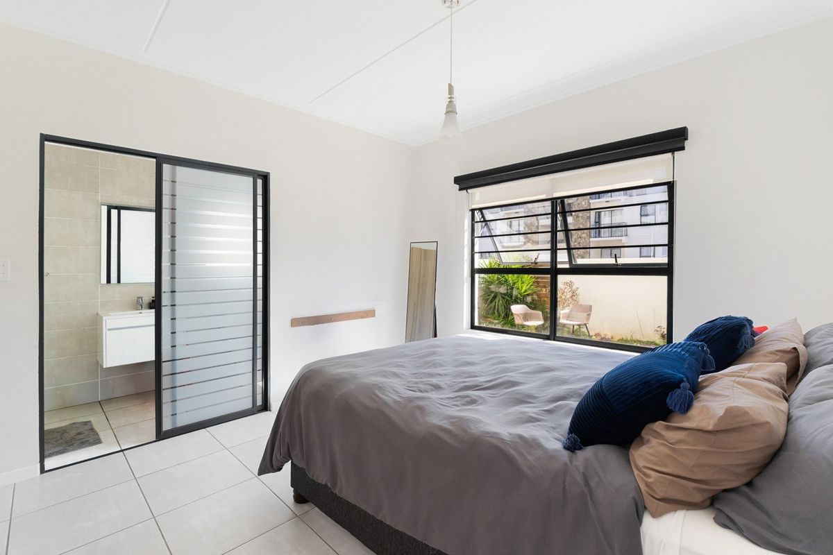 A bright bedroom featuring tiled flooring, a large window with black frames, and a modern en-suite bathroom accessed via a sliding glass door.