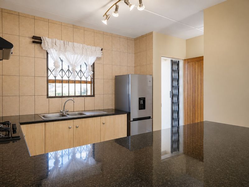 A functional kitchen featuring granite countertops, light wood cabinetry, a gas hob, and a tiled splashback, with a security gate leading to the exterior.
