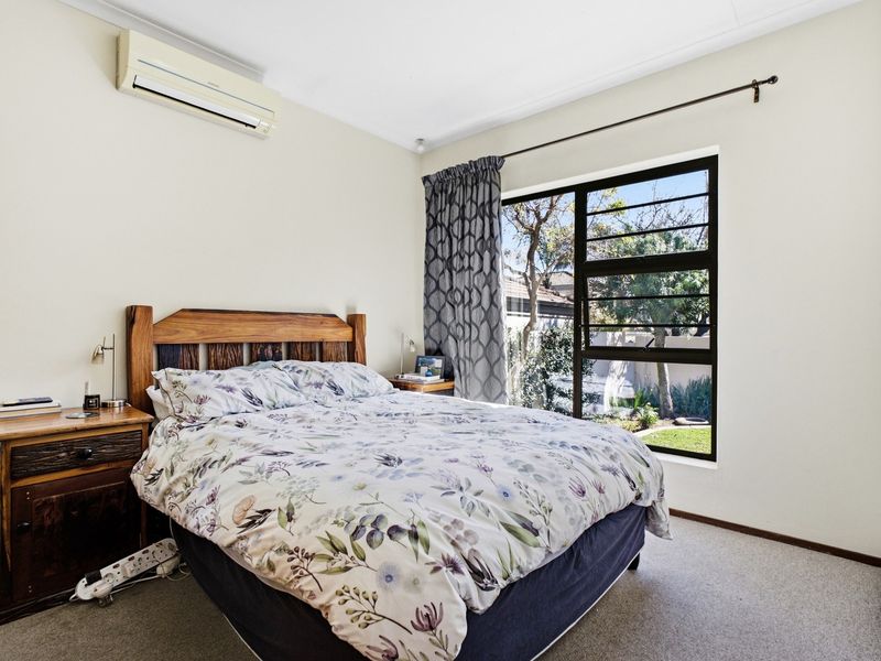A well-lit bedroom featuring neutral walls, carpeted flooring, and a split-system air conditioner, with a large window overlooking the garden.
