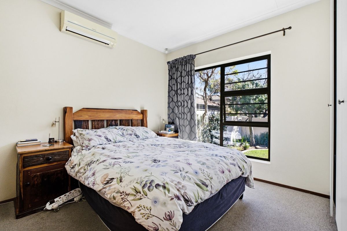 A well-lit bedroom featuring neutral walls, carpeted flooring, and a split-system air conditioner, with a large window overlooking the garden.