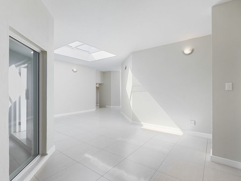 A bright, spacious, and empty living area featuring neutral tiled flooring, a large skylight, and modern wall-mounted lighting.