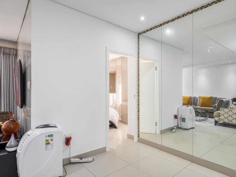A bright hallway featuring large wall-to-wall mirrors, polished tile flooring, and recessed lighting, providing access to adjacent living and bedroom areas.