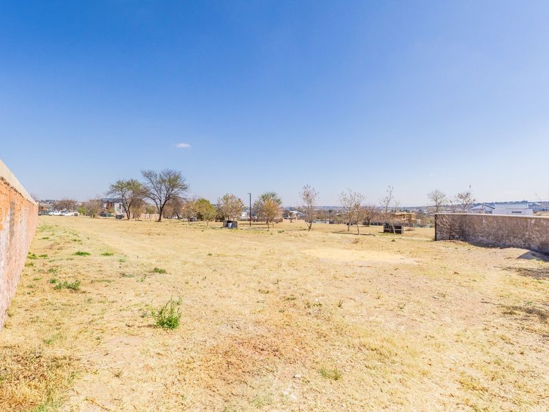 A vacant residential plot featuring dry grass, clear sky, and existing brick and concrete boundary walls.