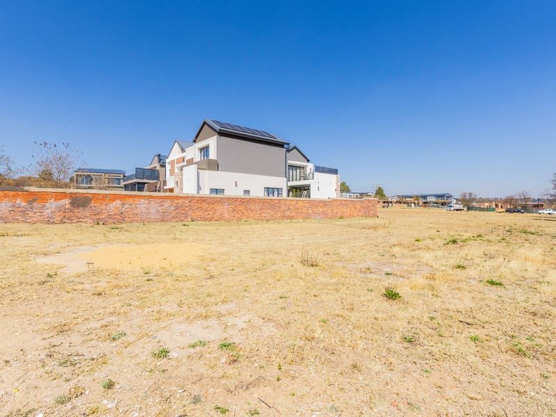 A vacant residential plot featuring a brick boundary wall with a view of neighboring modern multi-story homes under a clear blue sky.