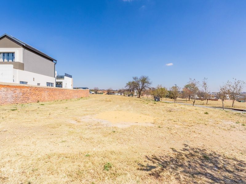 A vacant residential plot featuring dry grass, a brick boundary wall, and adjacent neighborhood views under a clear blue sky.