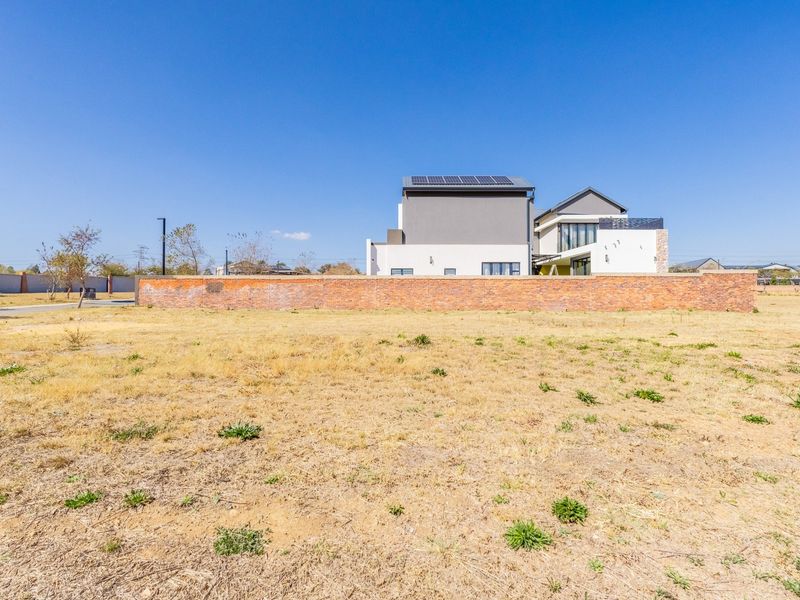 A wide-angle view of a vacant residential plot featuring a brick boundary wall and a modern multi-story home with solar panels in the background.
