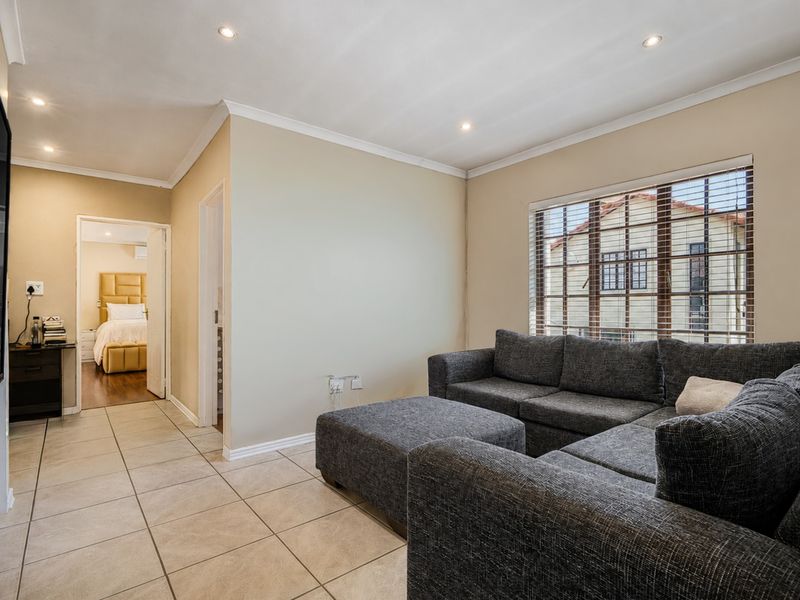 A well-maintained living room featuring neutral tiled flooring, recessed downlighting, and venetian blinds, with a view into an adjacent bedroom.