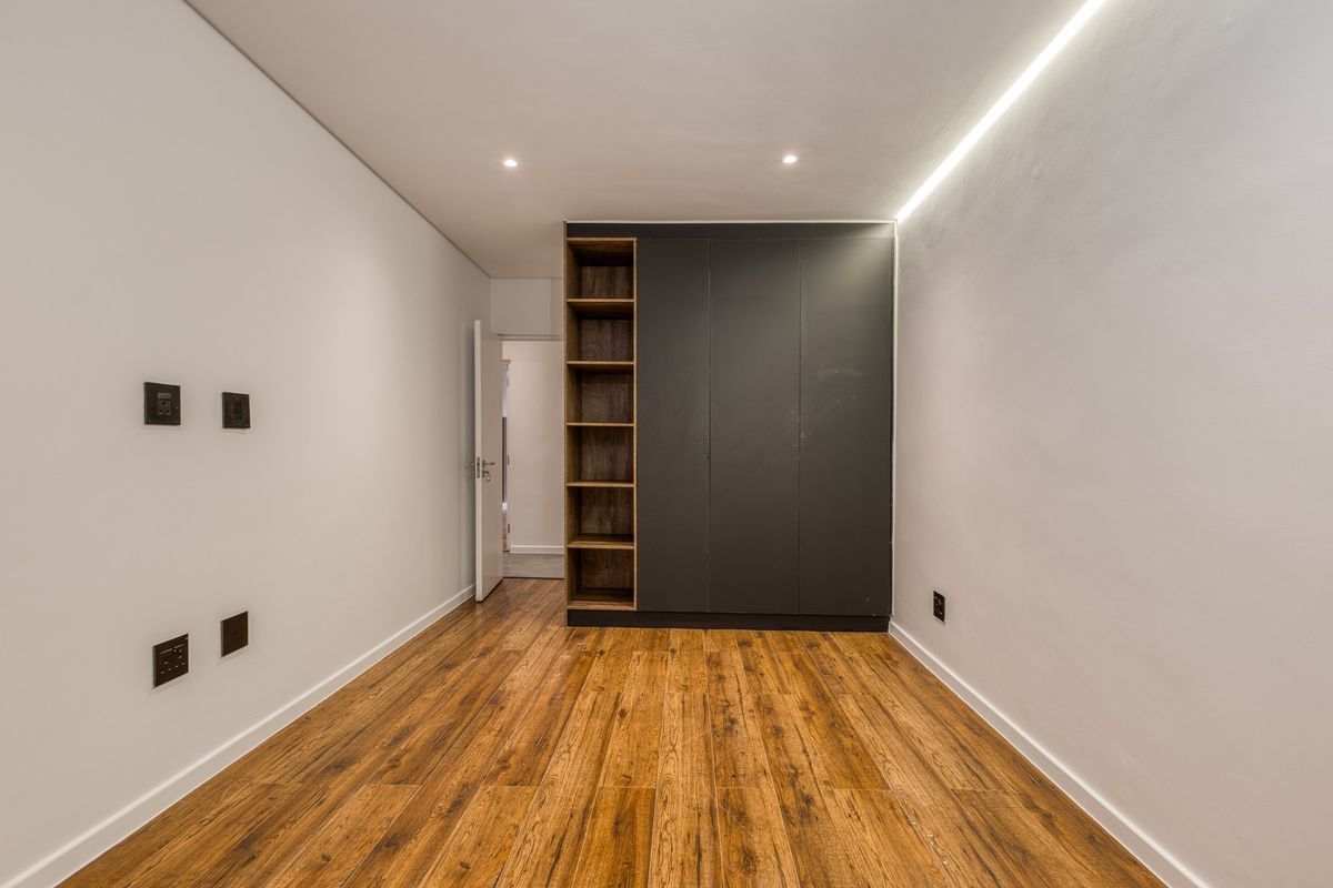 A modern, empty bedroom featuring dark built-in cabinetry with open shelving, warm laminate flooring, and integrated cove lighting.