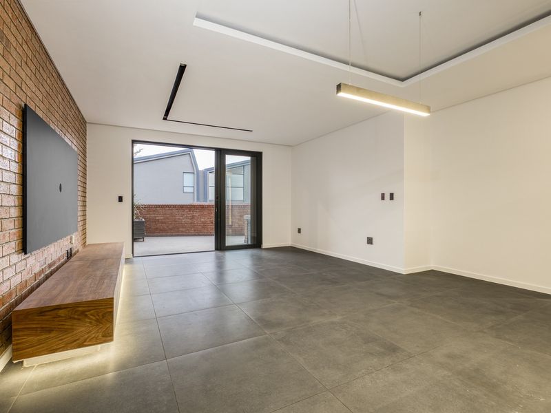 Modern open-plan living area featuring a striking exposed brick wall, large format floor tiles, recessed ceiling lighting, and direct access to an outdoor patio.