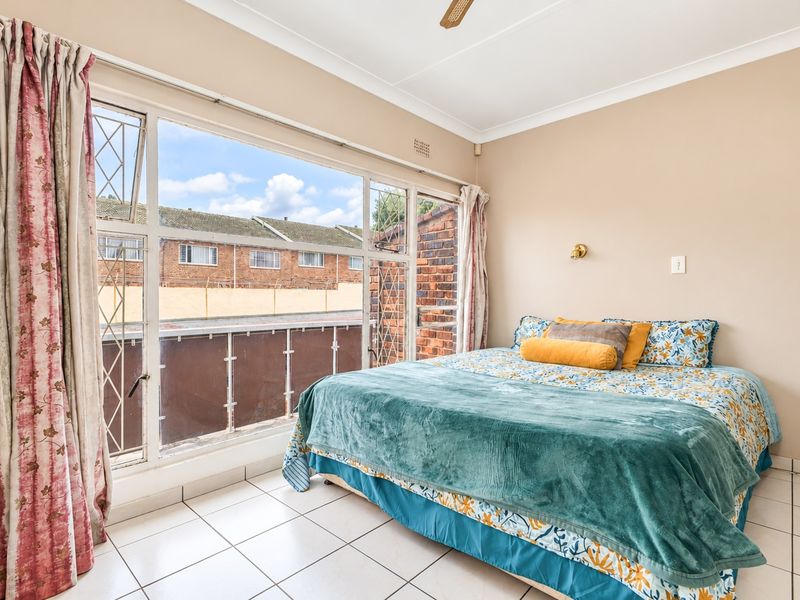 A bright bedroom featuring large windows with security bars, tiled flooring, and a ceiling fan, offering a functional space with natural light.