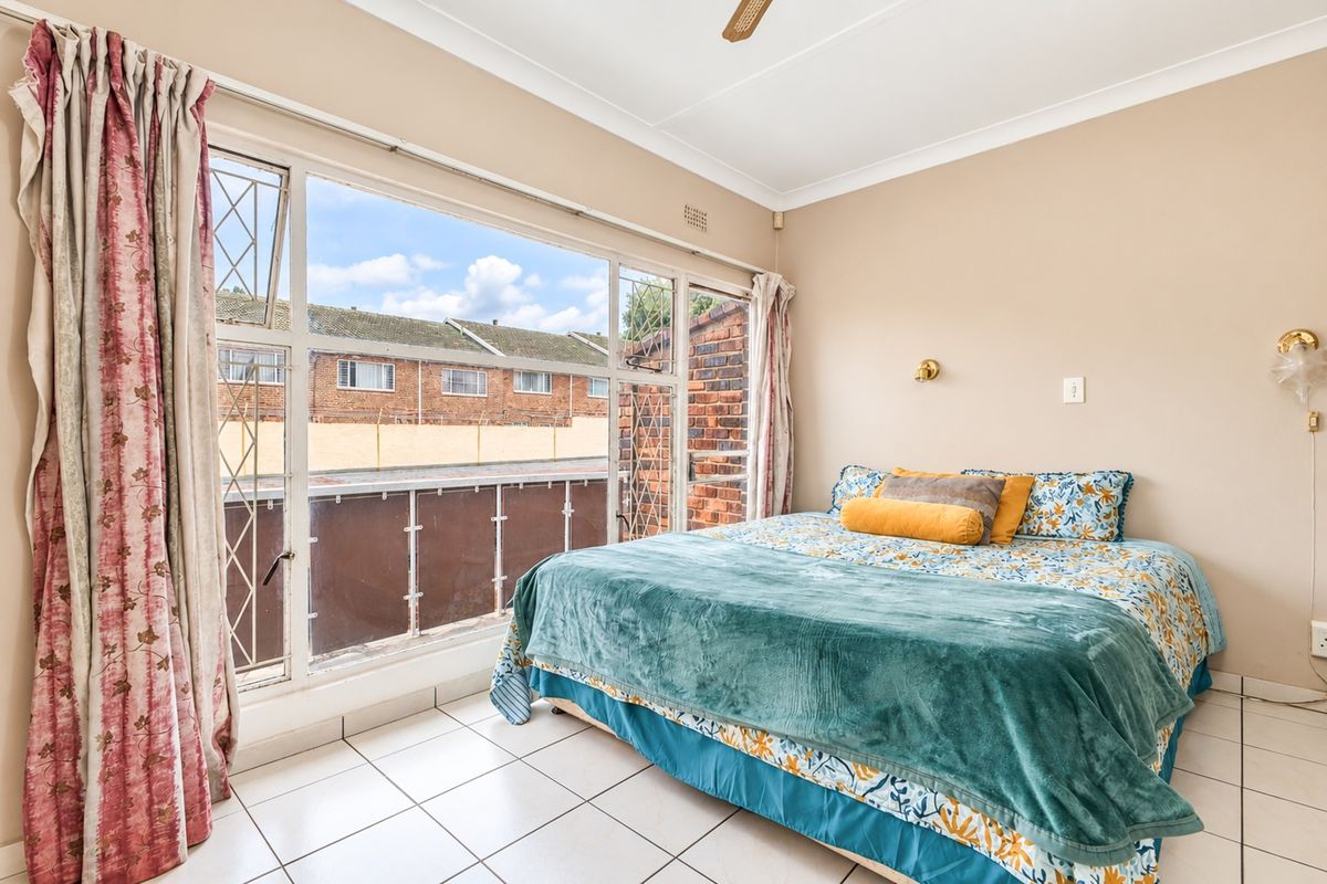A bright bedroom featuring large windows with security bars, tiled flooring, and a ceiling fan, offering a functional space with natural light.