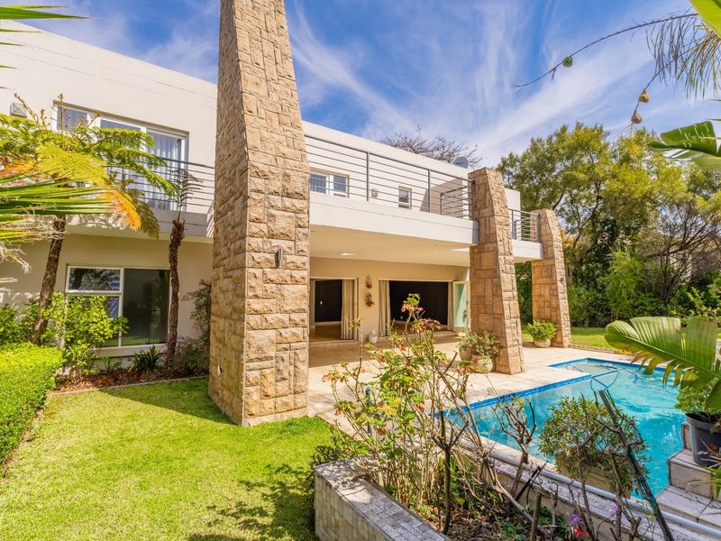 Rear view of a modern two-story home featuring stone-clad pillars, a spacious patio, an upper-level balcony, and a swimming pool set in a landscaped garden.
