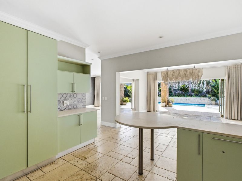 Modern kitchen featuring light green cabinetry, stone countertops, and travertine flooring, with an open-plan flow leading to a covered patio and swimming pool area.