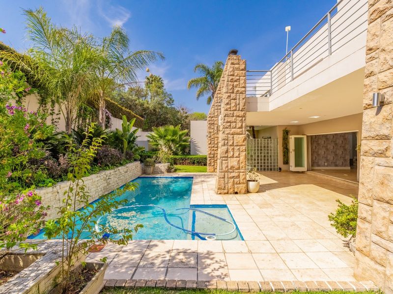 A well-maintained outdoor pool area featuring stone-clad pillars, a tiled patio, and a landscaped garden with a balcony above.