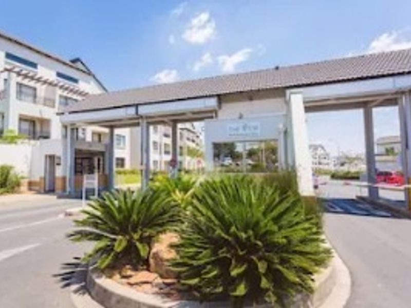 The main entrance and security gatehouse of the apartment complex, featuring a controlled access boom gate and well-maintained landscaped surroundings.