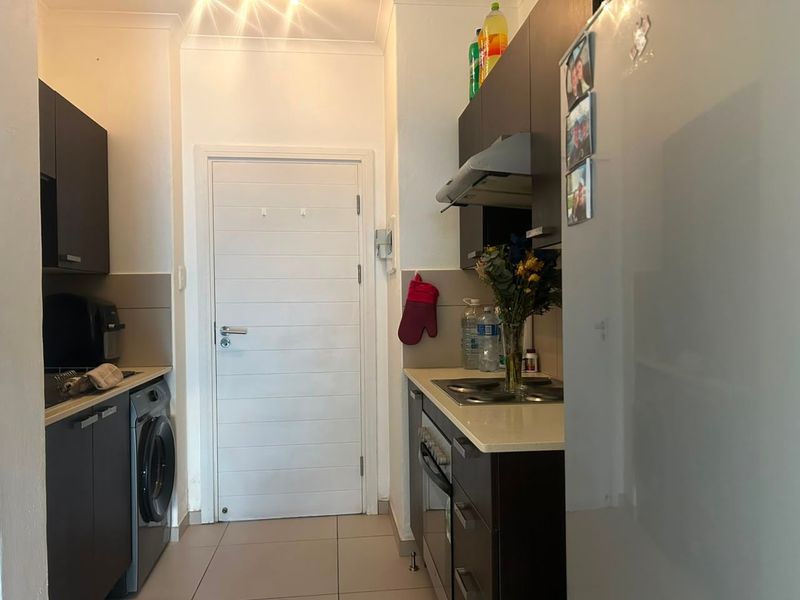 Compact galley-style kitchen featuring dark cabinetry, stone countertops, built-in oven, electric hob, extractor fan, and integrated laundry plumbing.