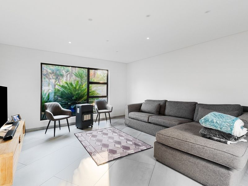 A bright and modern living room featuring large windows with garden views, recessed downlighting, and neutral tiled flooring.