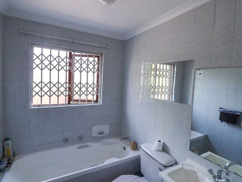 A functional bathroom featuring a bathtub, toilet, and vanity basin with tiled walls and a window fitted with a security gate.