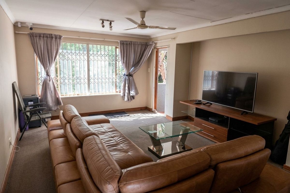 A living room featuring carpeted flooring, a ceiling fan, and security bars on the windows, providing access to an outdoor area.