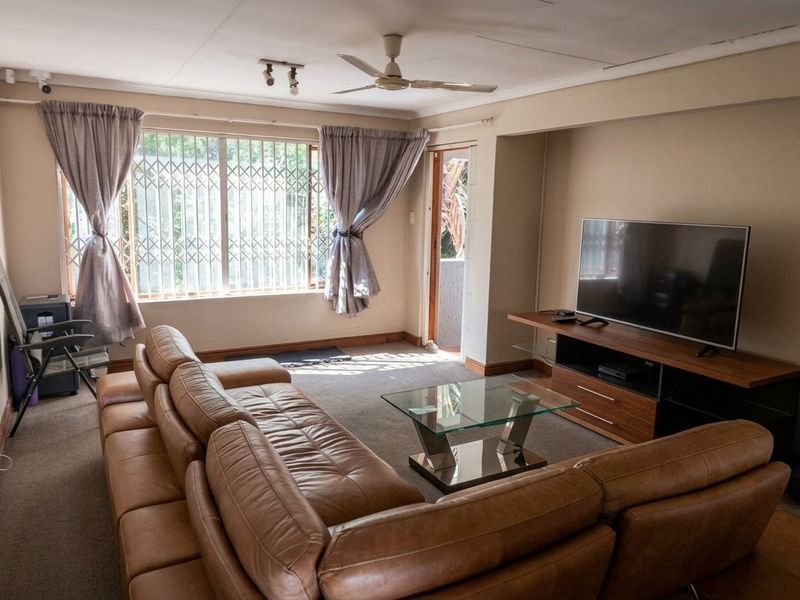 A living room featuring carpeted flooring, a ceiling fan, and security bars on the windows, providing access to an outdoor area.