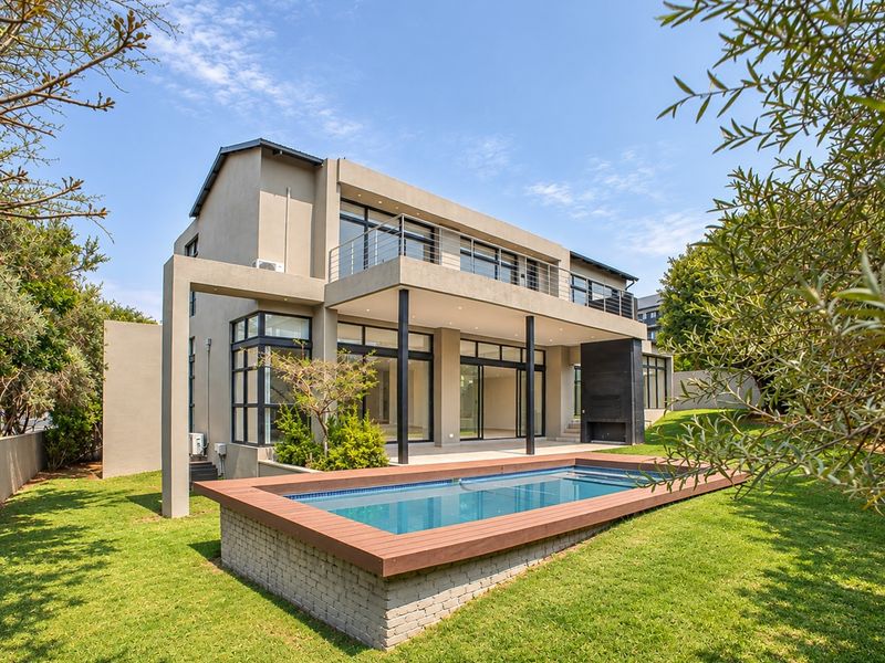 Rear view of a modern two-story house featuring a raised swimming pool, covered patio, balcony, and landscaped garden.