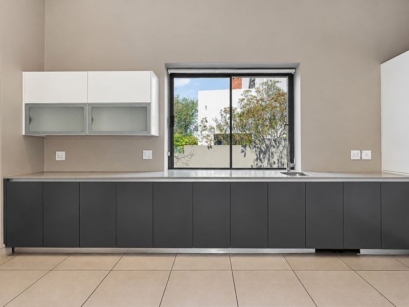 A modern, minimalist kitchen featuring sleek dark lower cabinetry, white upper cabinets, stone countertops, and a large sliding window overlooking the garden.