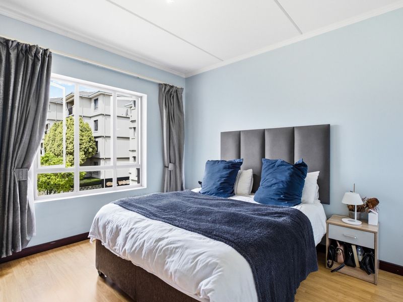 A well-lit bedroom featuring light-toned laminate flooring, neutral walls, and a large window providing natural light and an exterior view.